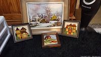 Four framed cross stitch pieces on black carpet: three small square frames and one large rectangular frame with village scene
