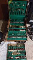 Four-tier wooden box with green felt lining open showing a complete set of flatware and serving utensils with wood handles and brass heads neatly arranged in rows.