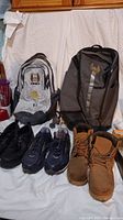 Photo shows four pairs of men's shoes and two backpacks arranged in front of a white backdrop. Shoes include black dress shoes, blue athletic shoes, brown boots, and black sneakers. Backpacks are gray with 'KINGS' logo and dark gray with eagle emblem.