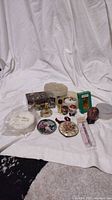 Overview of all lot items on white fabric showing several miniature perfume bottles, decorative boxes, plates, and powder puff