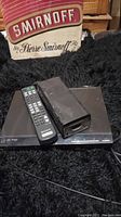 Front angle showing Sony DVD player with remote control and power adapter on black fuzzy surface with Smirnoff pillow in background