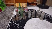 Full view of the 12 glass bottles arranged on a black and white rug. Various shapes and colors visible including labeled soda pop bottles and smaller medicine bottles.