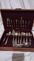 Open wooden box with burgundy velvet interior holding approximately 31 pieces of WB Rogers flatware including forks, knives, spoons, and serving utensils displayed upright and lying flat in compartments.