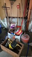Photo showing assortment of garden tools including rakes and hoe, wooden crate with yellow chainsaw, multiple gasoline cans, and boxed garden sprayer.