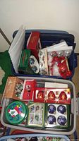 Tote filled with multiple boxes and loose Christmas decorations including bell ornaments and decorative tins.