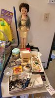 Photo showing vintage doll standing behind table with multiple costume jewelry pieces and trinket boxes arranged on it.