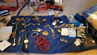 Wide photo showing assorted costume jewelry including multiple necklaces, bracelets, pins, watches, and packaging boxes on a blue cloth surface.