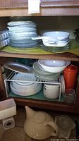Shelf with stacked white Corelle plates, glass plates, glass bowls, and Corning Ware pans with blue cornflower pattern.