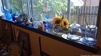 Wide view of glass items arranged on a window ledge including blue and clear glass pieces, a floral vase, and canisters, under natural daylight.