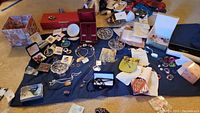 Overview of mixed jewelry items displayed on a dark cloth, including boxes, earrings, rings, necklaces, pins, jewelry bags, and a glass ring holder.