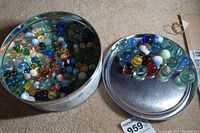 Wide shot of round metal tin filled with many glass marbles of assorted colors and designs, lid on the floor next to it showing some marbles against silver lid.