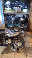 Wide view of various pots and pans stacked on shelves and floor including All-Clad, Cuisinart, Revere Ware, and Vollrath.