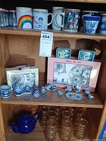 Full lot view showing two collectible tea sets displayed in boxes and arranged outside, blue ceramic teapot, and a row of gold-rimmed drinking glasses.