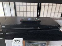 Front view of Panasonic Blu-ray player with remote on top, showing the USB port and model label.