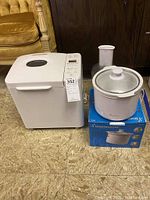 Breadman bread machine, white, with top window and control buttons alongside white slow cooker with glass lid on box