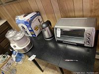Four small kitchen appliances including a white chocolate fountain, frozen yogurt maker, silver and black electric kettle, and Oster toaster oven placed on black table against wood panel wall.