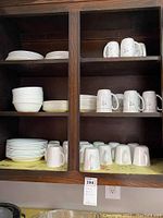 Wooden cabinet shelves with stacked white plates, bowls, and multiple white mugs with floral/script designs, arranged in neat rows.