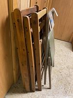 Photo showing side view of five vintage folding chairs leaning against wood-panel wall, including four wooden folding chairs and one green metal folding chair.
