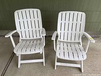 Front view of two white folding patio chairs with slatted backs and seats, wide armrests, shown folded upright and placed side by side.