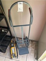 Metal hand truck standing upright with gray frame and red handles, attached lot tag visible.
