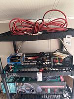 Three-tier metal rack with orange and black 50ft extension cord coiled on top. Sears battery charger black with red front label. Jumper cables with clamps beside charger. Craftsman Bushwacker 22 trimmer box on bottom shelf partially obscured. Items display some rust and wear.