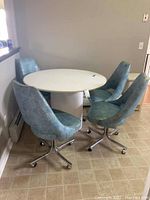 Front side view of four teal vinyl swivel chairs positioned around the white round pedestal kitchen table, showing chair design and table top.