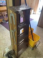 Wooden folding ladder with metal hinges and yellow mop bucket partially visible behind it