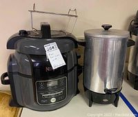 Front view showing the Ninja Foodi digital pressure cooker with control panel and metal rack, and adjacent metal coffee urn with spout and lid.