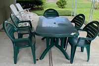 Photo showing one green plastic rectangular patio table surrounded by five matching green plastic armchairs on outdoor pavement.