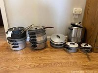 Full view of pots and pans in various sizes. Aluminum, stainless steel, and enamel pots visible with black handles, some with lids.