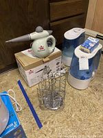 Photo displaying a Bissell Steam Shot handheld steamer with its box, two blue and white humidifiers, and a metal cylindrical rack on a floor.