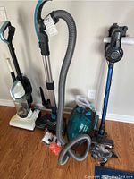 Photo showing all three vacuum cleaners standing upright along the wall, with the Jashen vacuum featuring a flexible hose and accessories laid out nearby.