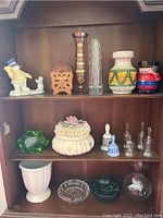 Shelf with various decorative items including a ceramic snowman, glass and ceramic vases, colorful nutcracker, wooden coasters, glass bells, lidded trinket dish and ashtrays.