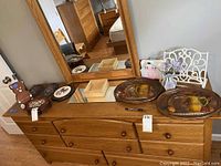 Various wall hangings and decor items displayed on a wooden dresser, including two painted wooden trays, jewelry box, mirror, and a white cast iron decorative stand.