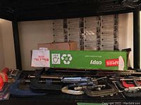 Photo of various bar clamps, C-clamps and other clamps on a shelf with boxes.