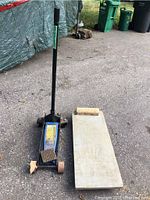 Photo of blue hydraulic floor car jack with black handle and wooden mechanic creeper outdoors with asphalt and green garbage bins in background.