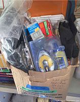 Cardboard box with various items including glue sticks, metal tool parts, Stanley glue stick, hardware tools wrapped in plastic bags.