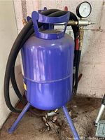 Blue metal 5 gallon pressure sand blaster with hose and pressure gauge, showing front view with operational label.