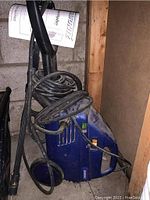 Overall view of blue Simoniz pressure washer with attached hose coiled on unit, showing wheels and handle for mobility.