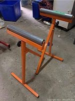 Photo shows two orange metal wood roller stands with cylindrical steel rollers on top, placed on a concrete floor in a workshop setting. The stands have adjustable height via knobs.