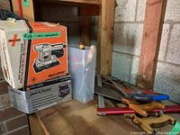Photo showing Black & Decker sanding tools boxed together with miscellaneous hand saws and tools on a wooden shelf in a garage area.