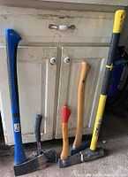 Sledgehammer with yellow handle, two axes with natural wood handles, one axe with blue handle leaning against white cabinet
