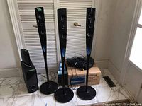 Three tall Logitech tower speakers and subwoofer beside large LG speaker/subwoofer with packaging for other items on floor