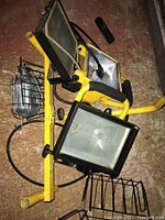 Three yellow industrial work floodlights resting on a textured floor. One has a metal protective cage, another has a black frame, cables attached, mounted on a yellow frame as a set.