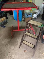 Full image showing red metal work table on caster wheels next to brown Ekco stepstool with three steps.