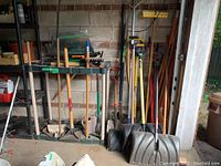 Corner view showing long-handled shovels, rakes, shovel scoops, tamper and broom leaning against the wall