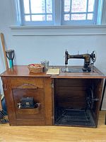 Antique Sovereign treadle sewing machine set into oak cabinet, opened door reveals accessory storage and hold flap, basket with sewing notions on cabinet top.