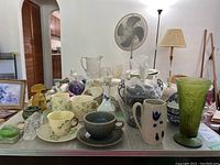 Wide view of the lot showing a variety of porcelain, glassware, and silverplate items displayed on a table with a lamp and fan in the background