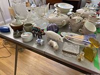 Wide table view of assorted items including Beswick ceramic pig figurines, Huichol beaded pig, various small dishes, perfume bottles, and beige and brown patterned china dishes with lids.