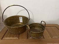 Photo showing the two brass pots on a wooden surface, larger pot with handle on left and smaller two-handled pot on right.